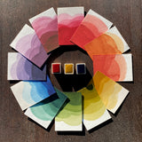 Ombre swatches of watercolour paint making up a colour wheel of Earthy colourway, surrounding Rose Madder, Yellow Ochre and Prussian Blue watercolour half pans, on a dark wooden background.