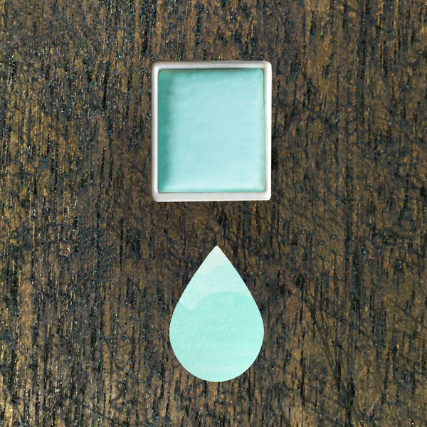 Aqua watercolour half pan above a droplet shaped ombre swatch in Aqua watercolour paint, on a rustic dark wooden background.