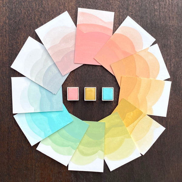 Ombre swatches of watercolour paint making up a colour wheel of the Grand Motel colourway, surrounding Blush, Naples Yellow and Aqua watercolour half pans, on a dark wooden background.