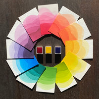 Ombre swatches of watercolour paint making up a colour wheel of Primary colourway, surrounding Permanent Rose, Yellow and Cerulean watercolour half pans, on a dark wooden background.