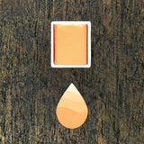 Apricot watercolour half pan above a droplet shaped ombre swatch in Apricot watercolour paint, on a rustic dark wooden background.