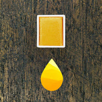 Golden Yellow watercolour half pan above a droplet shaped ombre swatch in Golden Yellow watercolour paint, on a rustic dark wooden background.